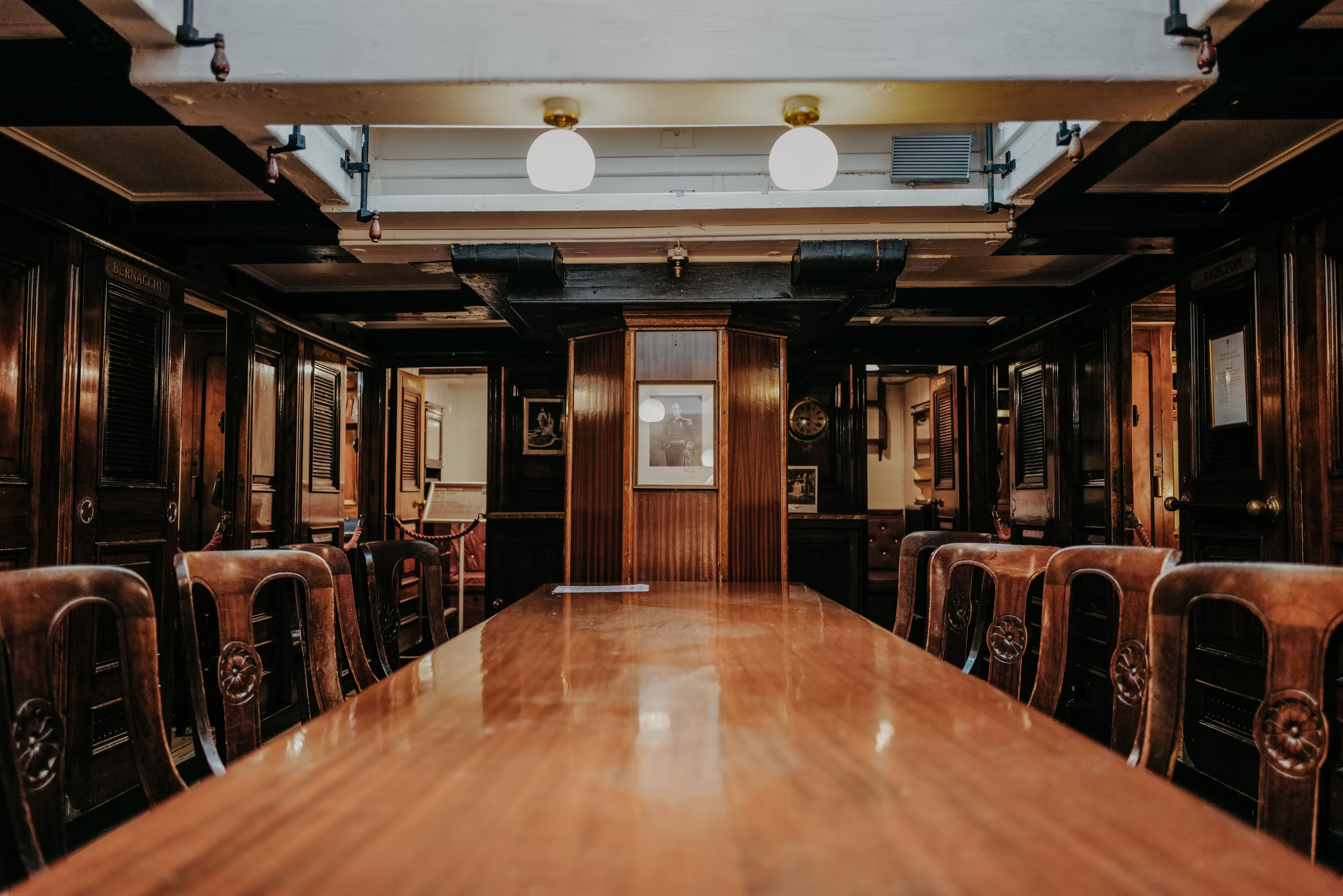 Looking down the mahogany table of RRS Discovery's wardroom. At the end of the table, a black and white portrait of Captain Scott is hung. Wooden chairs line each side of the table. Two round white lamps hang above from white overhead beams.