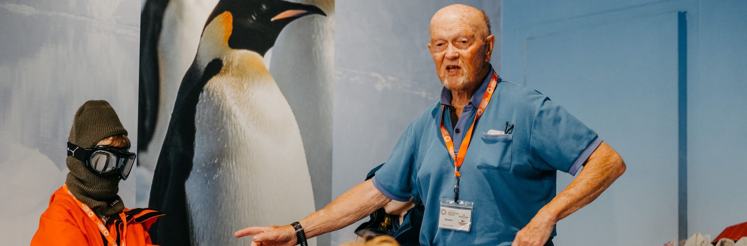 A volunteer wearing a bright orange Dundee Heritage Trust lanyard is talking with a group of children ages four to eight, who are listening attentively. One child is dressed as a polar explorer with balaclava and goggles! A penguin photograph is on the wall behind.